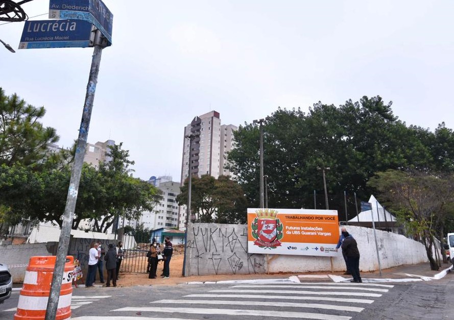 A foto mostra pessoas diante de um terreno grande de esquina com muro branco e portão aberto. Diante dele, à direita uma placa laranja anuncia futuras instalações da UBS, vemos a rua, a faixa de pedestres e uma placa à esquerda identificando rua Lucrécia